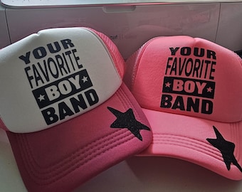 May include: Two trucker hats with the text "YOUR FAVORITE BOY BAND" in black. One hat is white and pink, the other is solid pink. Both have a black star on the side.