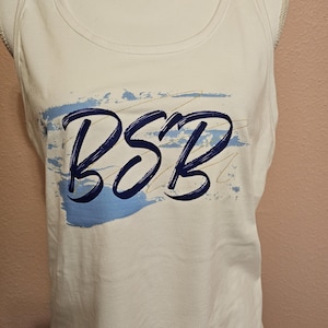 May include: White tank top with the letters "BSB" in dark blue script. The letters are over a light blue and gold brushstroke design. The tank top is displayed on a mannequin.