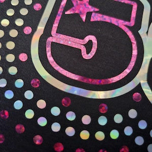 May include: Close-up of a black fabric with a pink and iridescent number 58 design. The number is outlined in a holographic material and features a star. Scattered iridescent and pink dots surround the number.