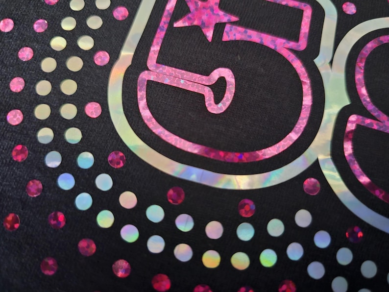 May include: Close-up of a black fabric with a pink and iridescent number 58 design. The number is outlined in a holographic material and features a star. Scattered iridescent and pink dots surround the number.