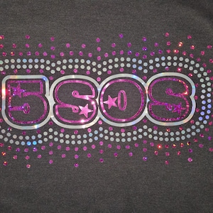 May include: Dark gray t-shirt with a sparkly design. The design features the letters "5SOS" in a pink and silver outline, with stars in the "O"s. The letters are surrounded by a dotted oval and scattered pink and silver sparkles.