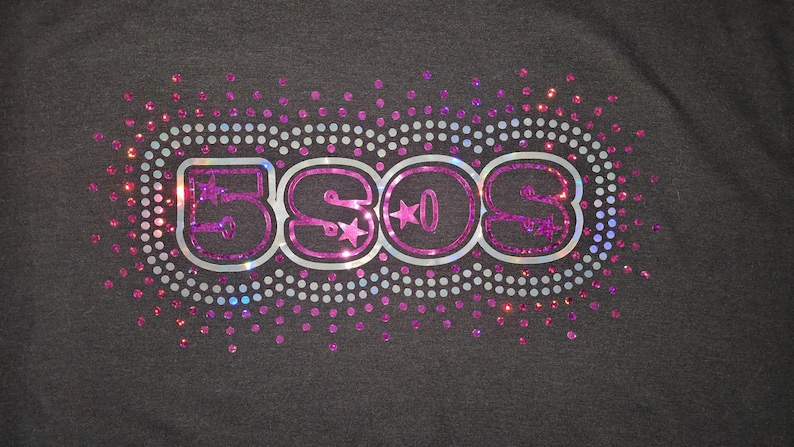 May include: Dark gray t-shirt with a sparkly design. The design features the letters "5SOS" in a pink and silver outline, with stars in the "O"s. The letters are surrounded by a dotted oval and scattered pink and silver sparkles.