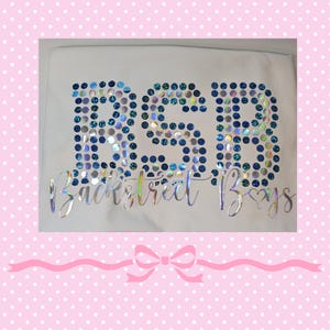 May include: A white t-shirt featuring the letters BSB formed by blue and iridescent circular sequins. Below the letters, the text Backstreet Boys is printed in a silver holographic script font with a small heart integrated into the letter y.