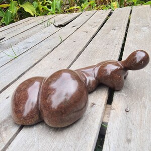 May include: A brown, smooth, abstract sculpture of a reclining figure with a rounded, bulbous shape. The sculpture is made of stone and has a glossy finish. It is resting on a wooden surface.