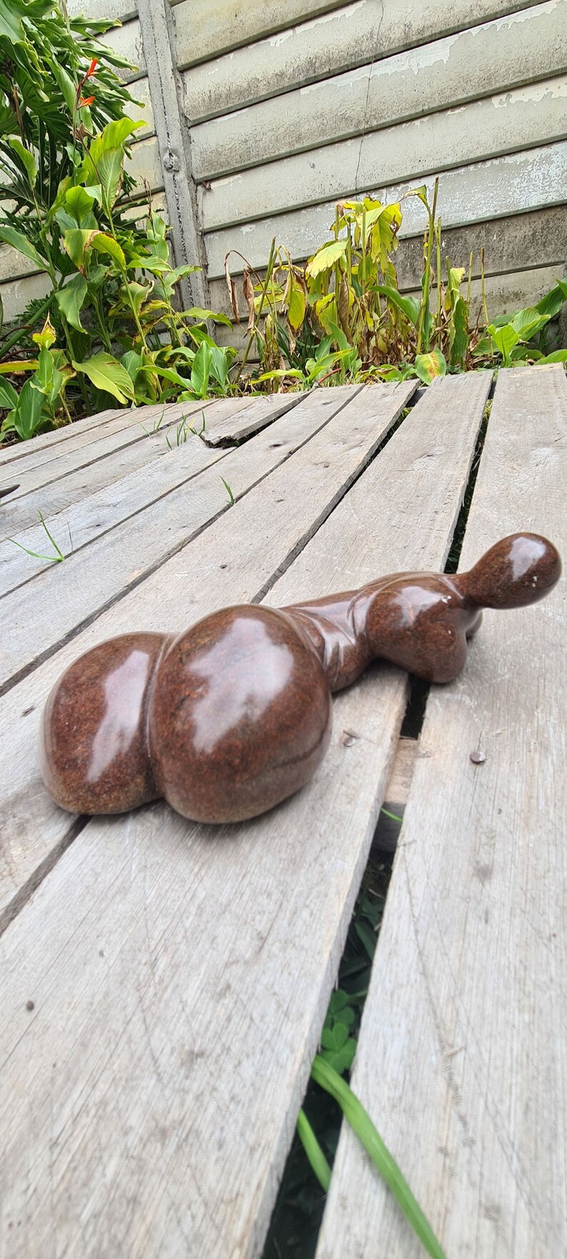 May include: A brown, smooth, abstract sculpture of a reclining figure with a rounded, bulbous shape. The sculpture is made of stone and has a glossy finish. It is resting on a wooden surface.