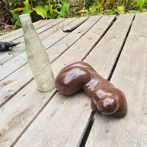 May include: A brown, smooth, abstract sculpture of a human figure lying on a wooden surface. A clear glass bottle is also on the surface.