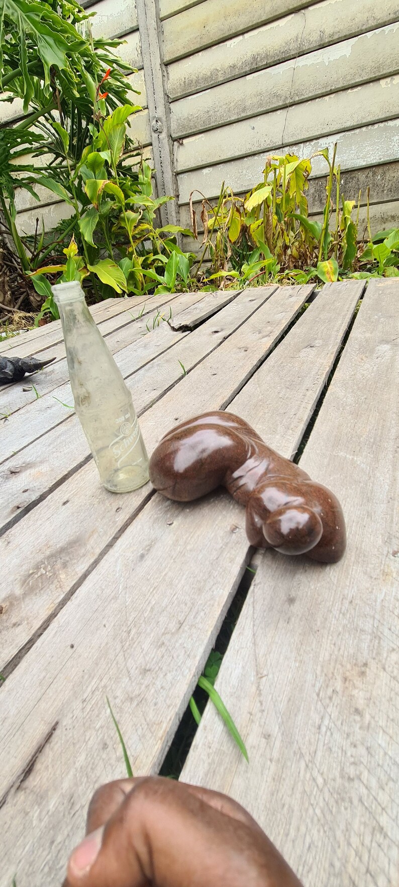 May include: A brown, smooth, abstract sculpture of a human figure lying on a wooden surface. A clear glass bottle is also on the surface.