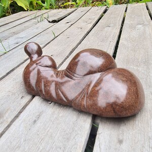 May include: A smooth, brown, abstract sculpture of a reclining figure. The sculpture is made of stone and has a polished finish. It is resting on a wooden deck.