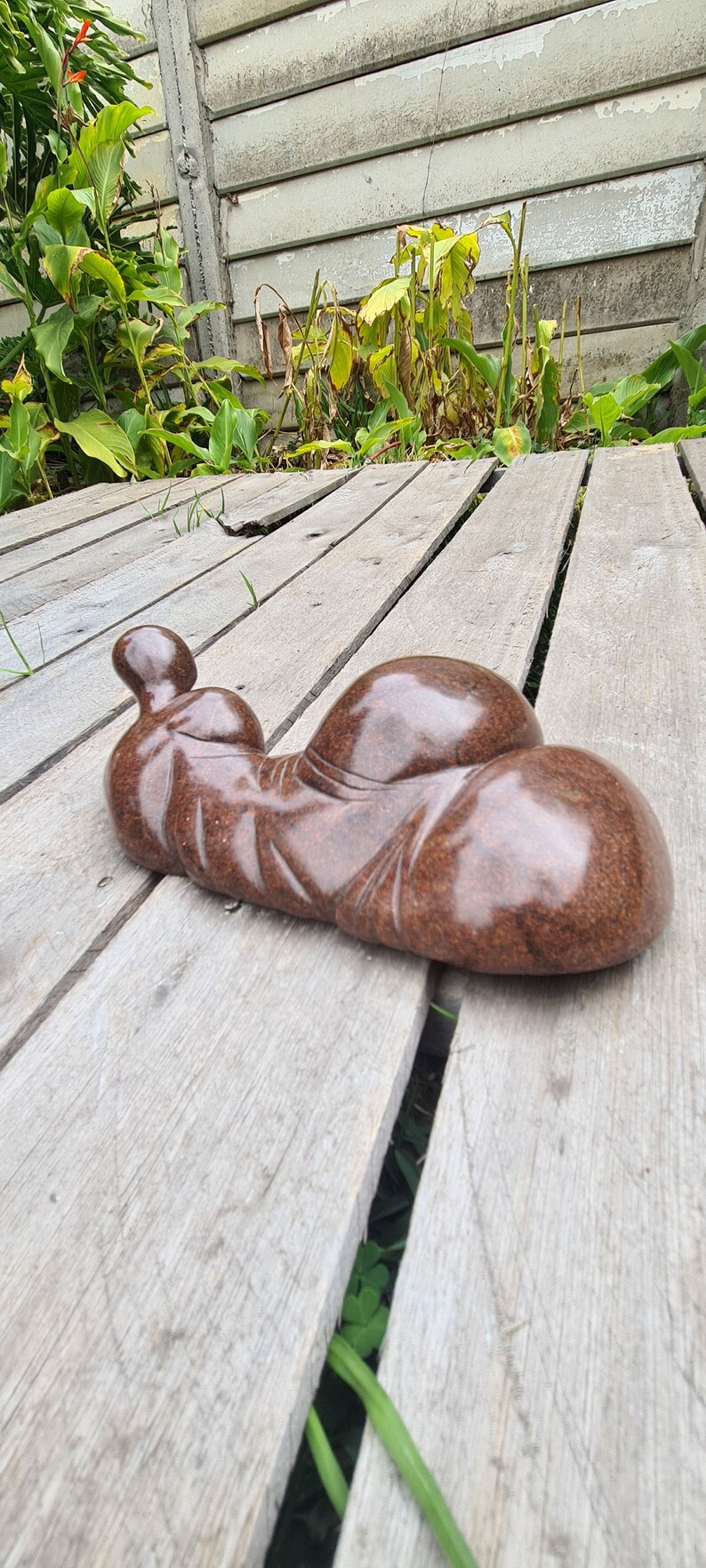May include: A smooth, brown, abstract sculpture of a reclining figure. The sculpture is made of stone and has a polished finish. It is resting on a wooden deck.