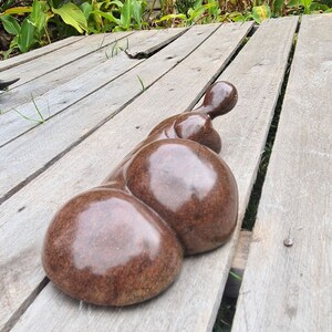 May include: A smooth, brown, abstract sculpture made of stone, resting on a wooden surface.