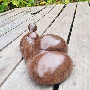 May include: A brown, abstract sculpture of a person lying down on a wooden deck. The sculpture has a smooth, polished surface and is made of a material that looks like stone.