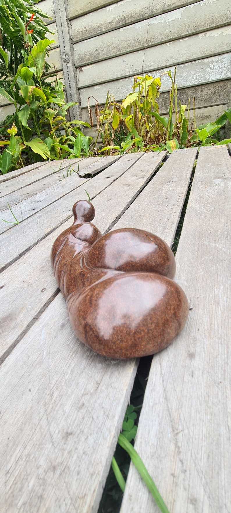 May include: A brown, abstract sculpture of a person lying down on a wooden deck. The sculpture has a smooth, polished surface and is made of a material that looks like stone.