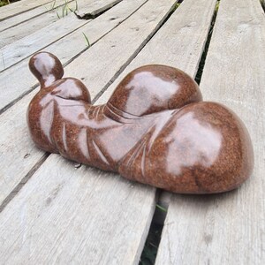 May include: A brown, smooth, abstract sculpture of a reclining figure. The sculpture is made of stone and has a polished finish. It is resting on a wooden deck.