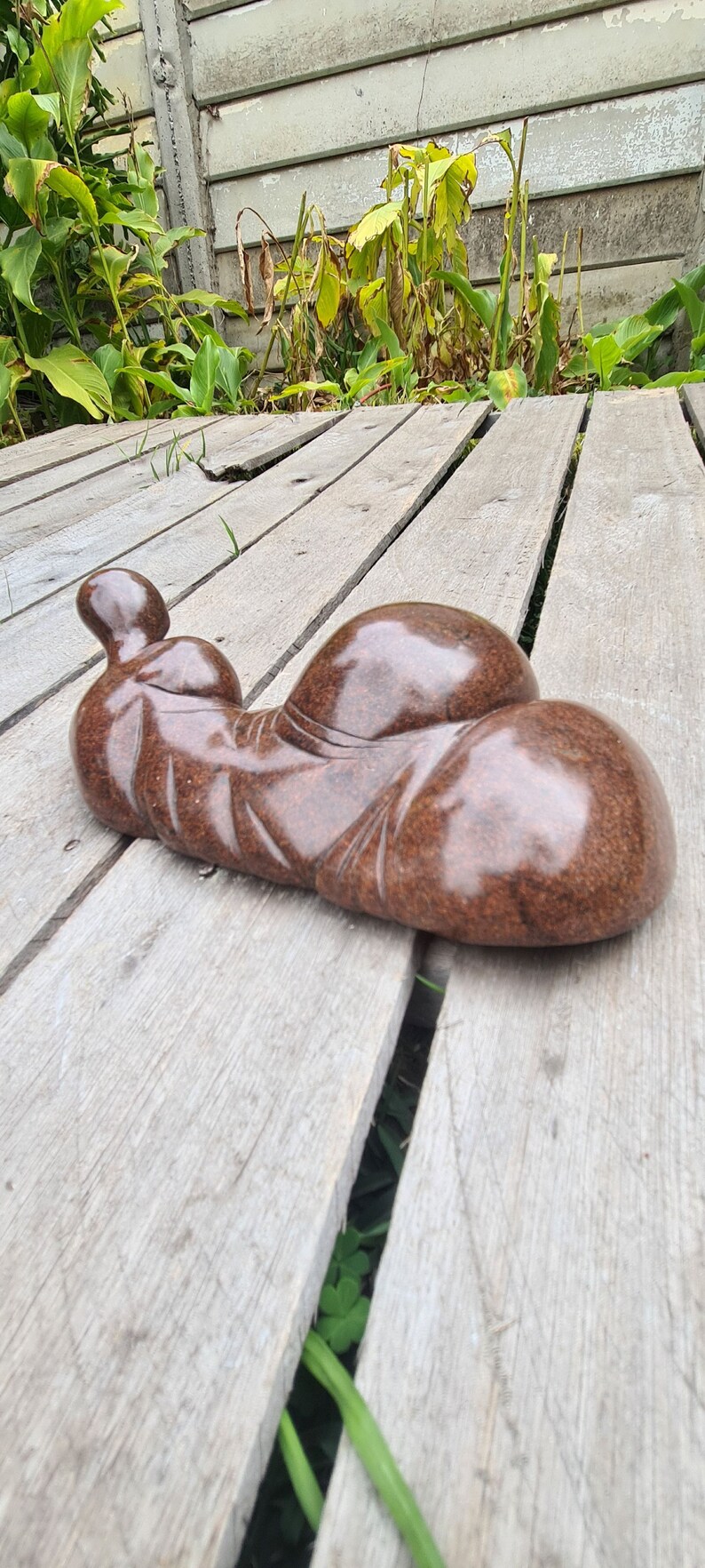 May include: A brown, smooth, abstract sculpture of a reclining figure. The sculpture is made of stone and has a polished finish. It is resting on a wooden deck.
