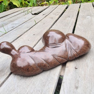 May include: A brown, abstract sculpture made of stone, with a smooth, polished finish. The sculpture is shaped like a reclining figure, with rounded forms and a flowing, organic design. It is resting on a wooden deck.