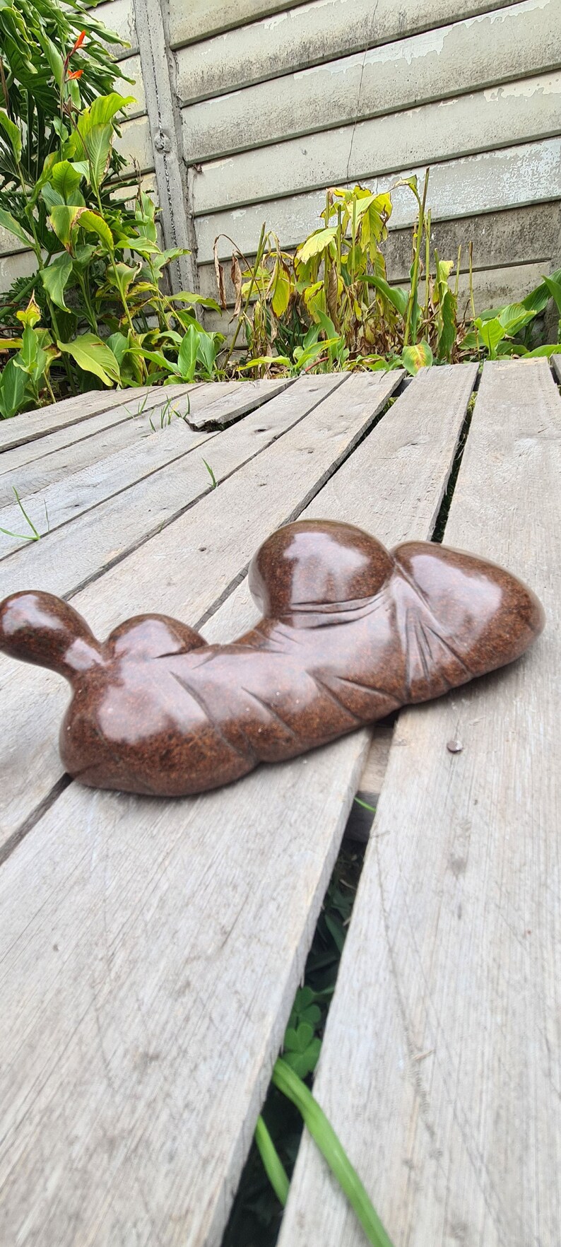 May include: A brown, abstract sculpture made of stone, with a smooth, polished finish. The sculpture is shaped like a reclining figure, with rounded forms and a flowing, organic design. It is resting on a wooden deck.