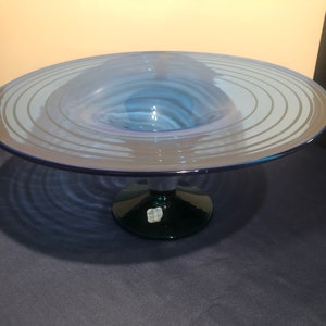 May include: A blue glass bowl with a wide, flat base and a deep, circular center. The bowl has a series of concentric rings around the center, creating a ripple effect. The bowl is on a green pedestal.