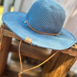 Könnte beinhalten: Ein blauer Sonnenhut mit breiter Krempe und einem braunen Lederband. Der Hut hat eine gewebte Textur und einen Kinnriemen aus Leder. Ein kleines Lederetikett ist am Hut befestigt.