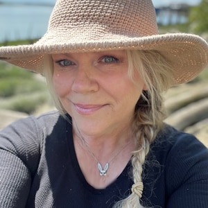 Peut inclure: Une femme aux cheveux blonds portant un chapeau beige à larges bords et une chemise noire à manches longues. Elle porte un collier en argent avec un pendentif en forme de lapin.