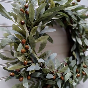 May include: A decorative wreath featuring olive branches and brown berries. The leaves are various shades of green and gray, creating a natural and rustic aesthetic. The wreath is likely used for home decor, adding a touch of nature to any space.