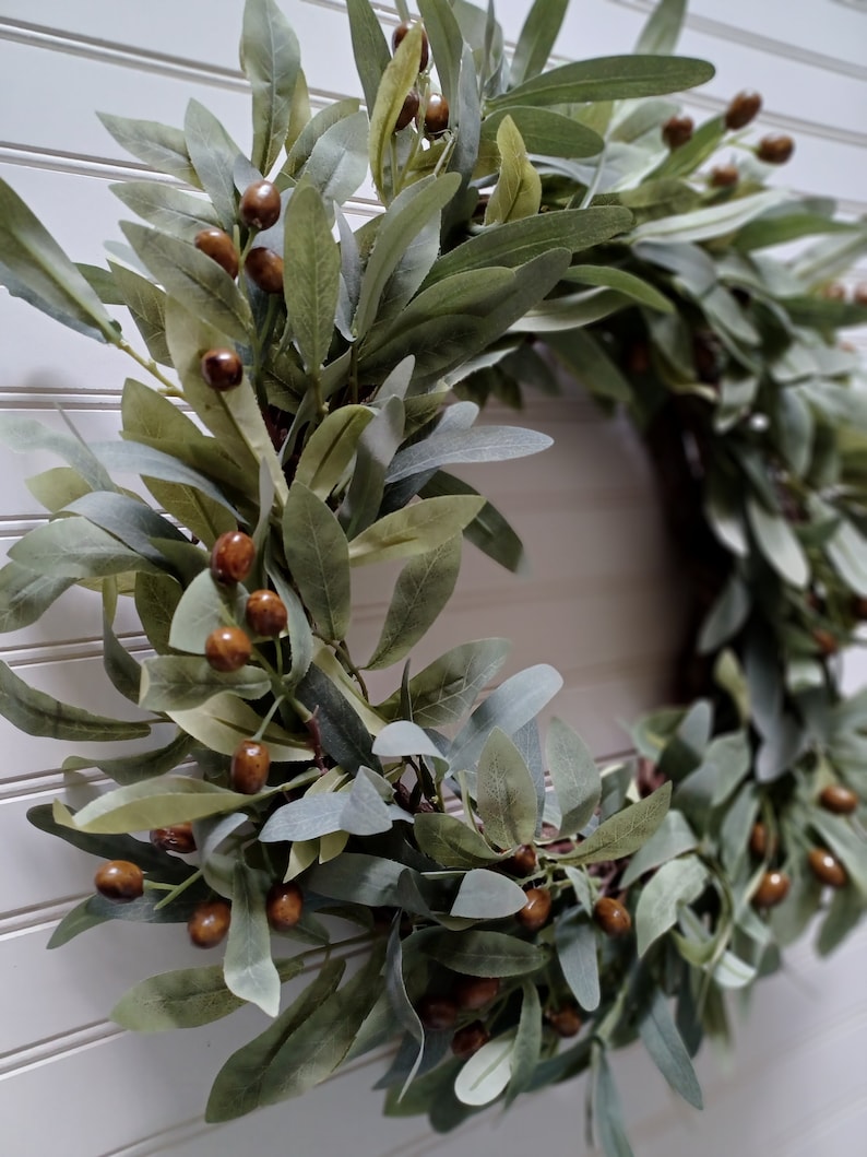 May include: A decorative wreath featuring olive branches and brown berries. The leaves are various shades of green and gray, creating a natural and rustic aesthetic. The wreath is likely used for home decor, adding a touch of nature to any space.