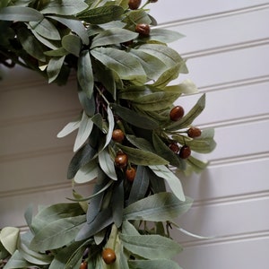 May include: A close-up of a decorative wreath featuring green leaves and brown berries. The wreath is set against a white background with vertical paneling. The leaves have a soft, muted green color.