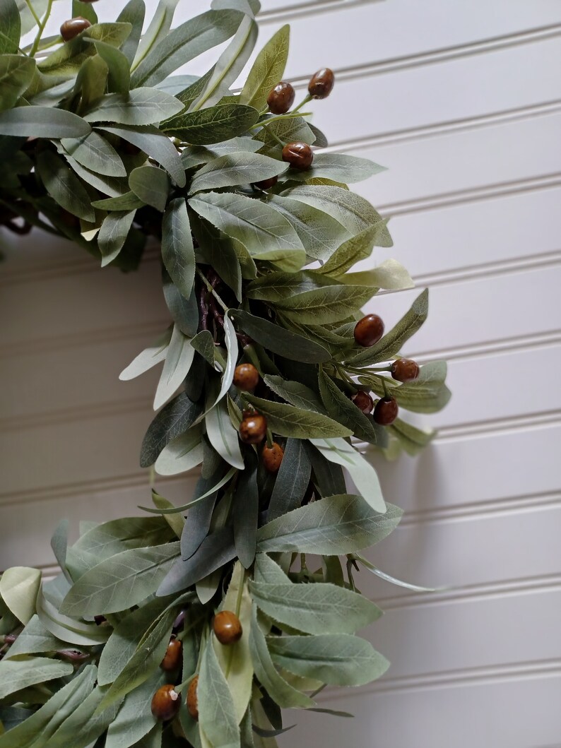 May include: A close-up of a decorative wreath featuring green leaves and brown berries. The wreath is set against a white background with vertical paneling. The leaves have a soft, muted green color.