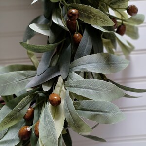 May include: Close-up of a decorative garland featuring faux olive branches and brown, round berries. The leaves are a muted green with a soft, textured appearance. The berries have a speckled, natural look, adding a touch of rustic charm to the design.
