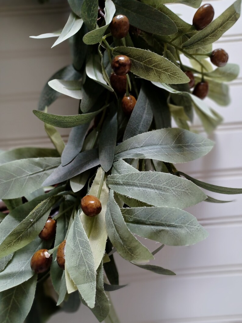 May include: Close-up of a decorative garland featuring faux olive branches and brown, round berries. The leaves are a muted green with a soft, textured appearance. The berries have a speckled, natural look, adding a touch of rustic charm to the design.