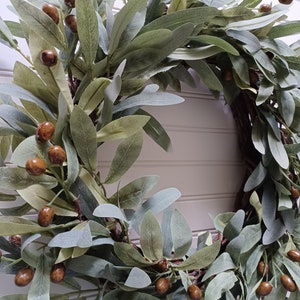 May include: A decorative wreath featuring olive branches with green and gray leaves and brown olive-like berries. The wreath is designed for home decor and adds a natural, rustic touch to any space.