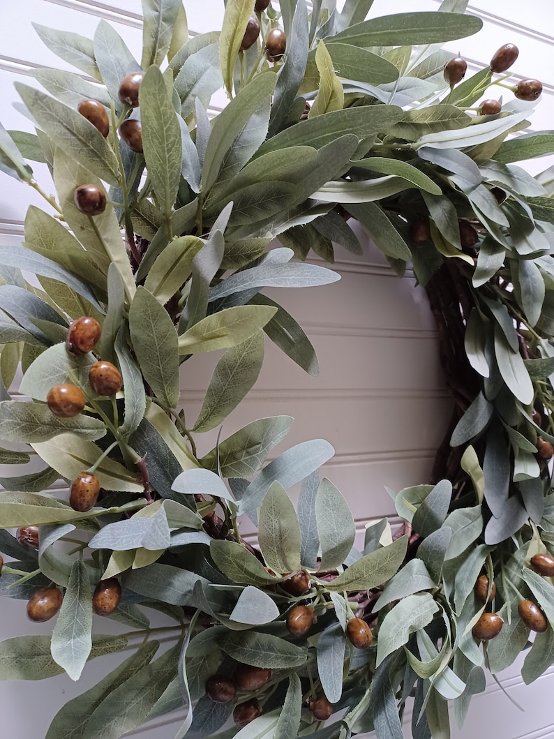 May include: A decorative wreath featuring olive branches with green and gray leaves and brown olive-like berries. The wreath is designed for home decor and adds a natural, rustic touch to any space.