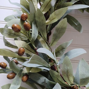 May include: Close-up of a decorative wreath featuring olive branches and brown, olive-shaped accents. The leaves are a mix of green and gray-green, creating a natural, rustic aesthetic. The wreath is suitable for home decor.