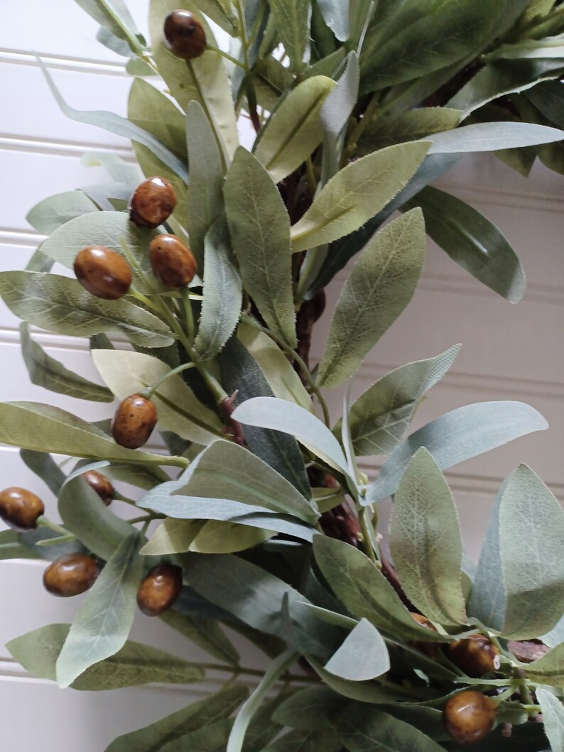 May include: Close-up of a decorative wreath featuring olive branches and brown, olive-shaped accents. The leaves are a mix of green and gray-green, creating a natural, rustic aesthetic. The wreath is suitable for home decor.