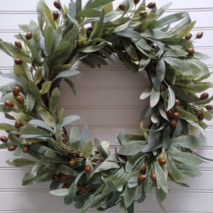 May include: A circular wreath featuring green and gray-green leaves and small brown berries. The wreath is made of interwoven branches and is displayed against a white, slatted background. This decorative wreath is suitable for indoor or outdoor use.