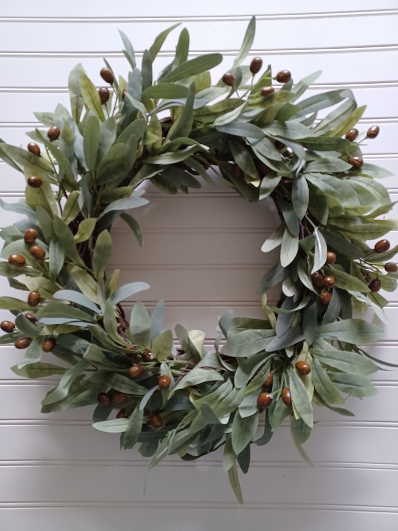 May include: A circular wreath featuring green and gray-green leaves and small brown berries. The wreath is made of interwoven branches and is displayed against a white, slatted background. This decorative wreath is suitable for indoor or outdoor use.