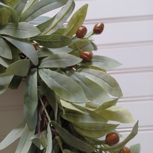 May include: Close-up of a decorative wreath featuring olive green leaves and brown berries. The leaves have a soft, muted green color, and the berries are a rich, warm brown. The wreath is set against a white background.