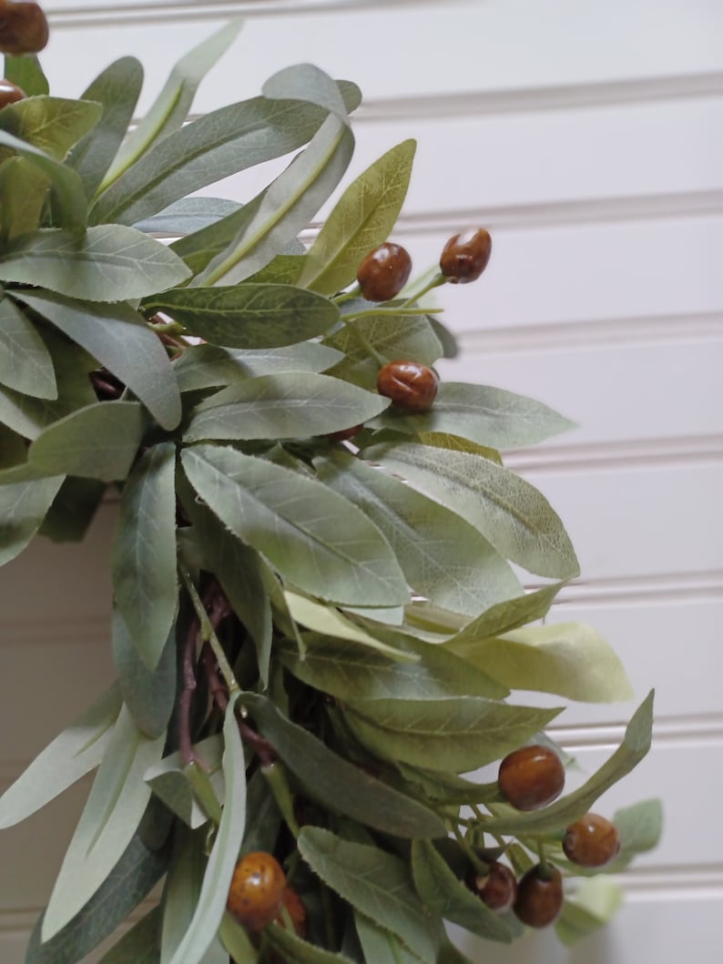 May include: Close-up of a decorative wreath featuring olive green leaves and brown berries. The leaves have a soft, muted green color, and the berries are a rich, warm brown. The wreath is set against a white background.