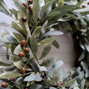 May include: A close-up of a decorative wreath featuring olive branches and brown olive-like berries. The leaves are various shades of green and gray, creating a natural and rustic aesthetic. The wreath is designed for home decor.