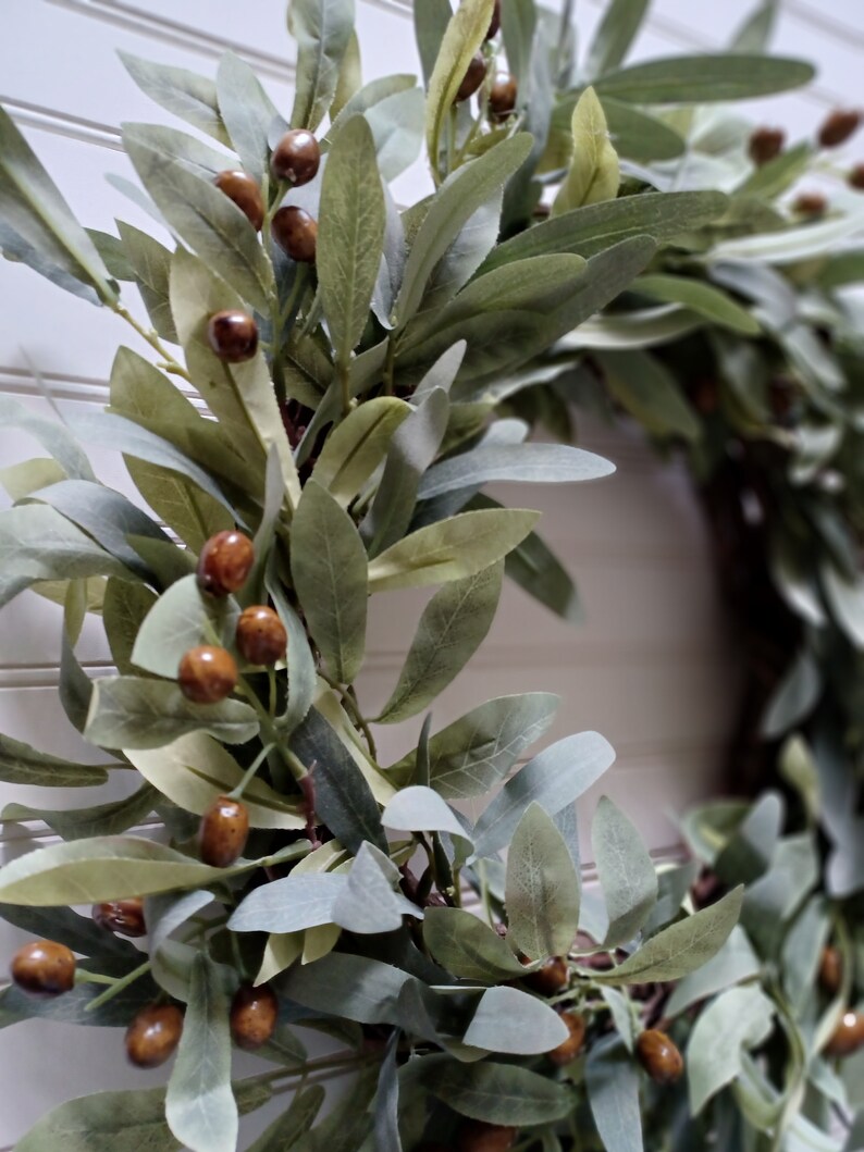 May include: A close-up of a decorative wreath featuring olive branches and brown olive-like berries. The leaves are various shades of green and gray, creating a natural and rustic aesthetic. The wreath is designed for home decor.