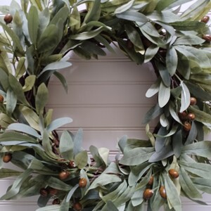 May include: A circular wreath featuring olive green leaves and small brown berries. The wreath is made of natural materials and is suitable for home decor. The leaves have a soft, muted green color, and the berries add a touch of warmth.