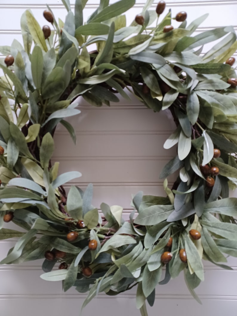 May include: A circular wreath featuring olive green leaves and small brown berries. The wreath is made of natural materials and is suitable for home decor. The leaves have a soft, muted green color, and the berries add a touch of warmth.