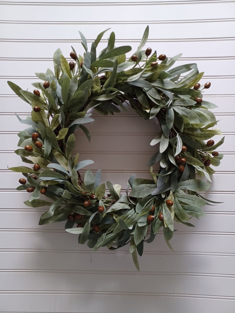 May include: A circular wreath featuring green leaves and small brown berries. The leaves have a muted green color, and the berries are a deep brown. The wreath is a decorative item, suitable for seasonal or year-round display.