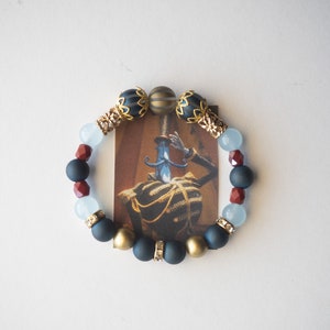 May include: A beaded bracelet featuring a variety of beads in navy, gold, red, and light blue. The bracelet is centered around a small card with a vintage illustration of a person in a military uniform.