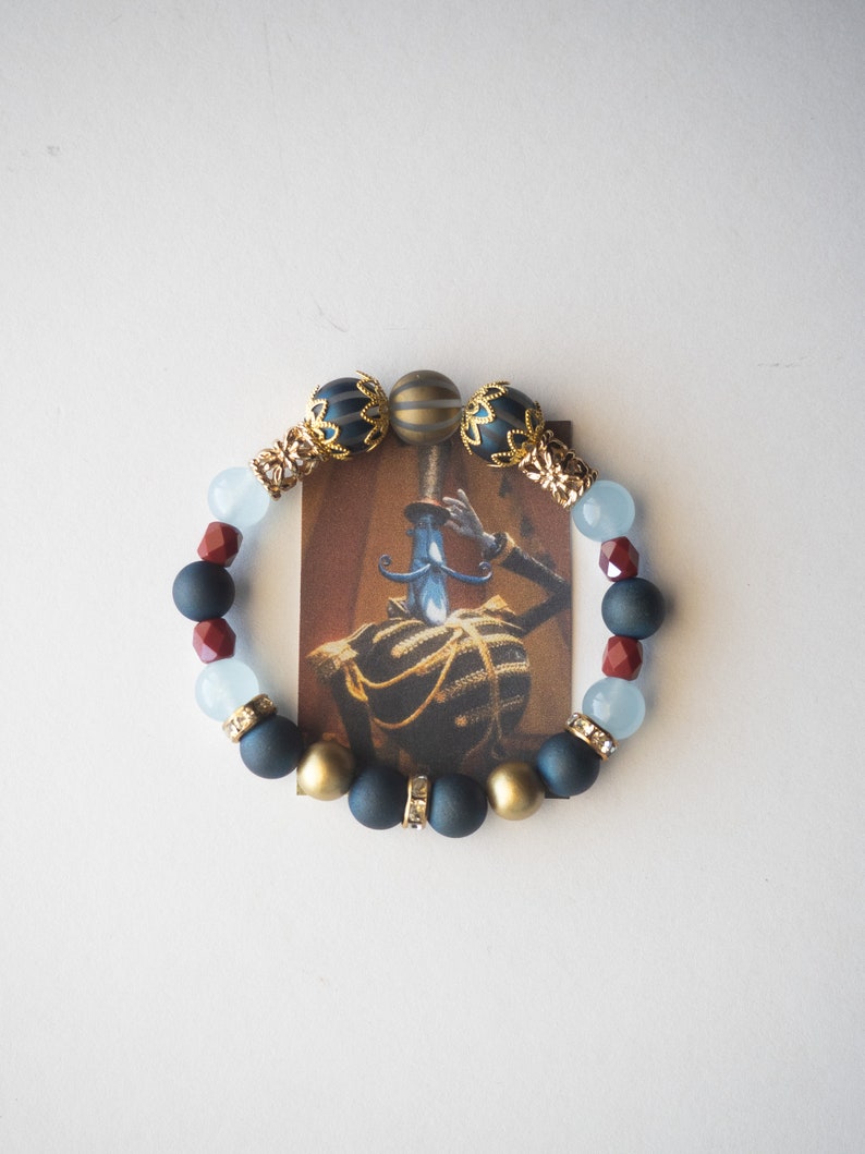 May include: A beaded bracelet featuring a variety of beads in navy, gold, red, and light blue. The bracelet is centered around a small card with a vintage illustration of a person in a military uniform.