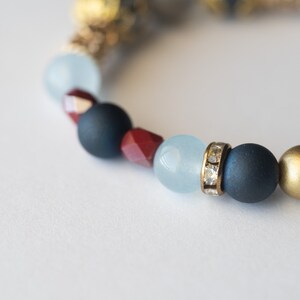 May include: A beaded bracelet featuring a mix of colors and textures. The bracelet includes navy blue, red, light blue, and gold beads. Some beads are matte, while others have a metallic or crystal-like finish. The bracelet is on a white background.