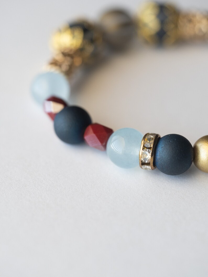 May include: A beaded bracelet featuring a mix of colors and textures. The bracelet includes navy blue, red, light blue, and gold beads. Some beads are matte, while others have a metallic or crystal-like finish. The bracelet is on a white background.