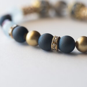 May include: A close-up of a beaded bracelet featuring matte navy blue, gold, and clear beads. Some beads are accented with small, clear crystals. The bracelet is on a white background.