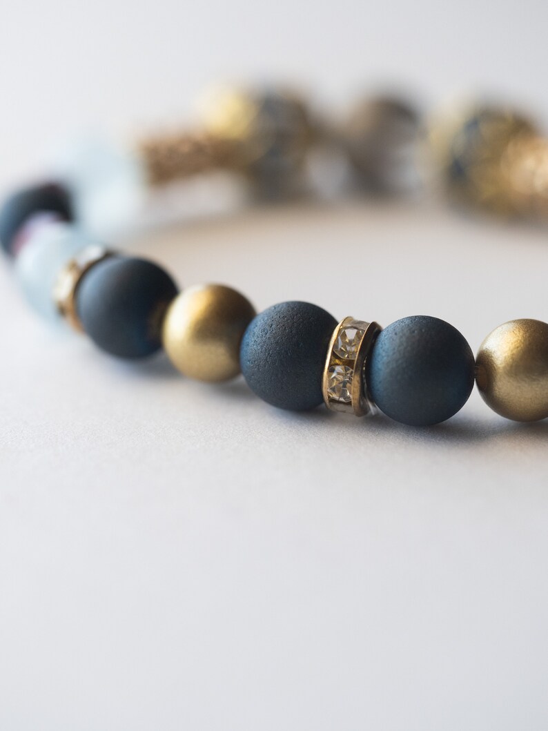 May include: A close-up of a beaded bracelet featuring matte navy blue, gold, and clear beads. Some beads are accented with small, clear crystals. The bracelet is on a white background.