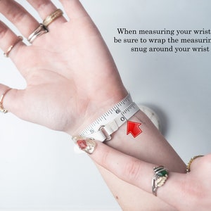 May include: A white measuring tape wrapped around a wrist, indicating a wrist size of approximately 6 inches. The image includes text advising to wrap the tape snugly. Several rings adorn the fingers.
