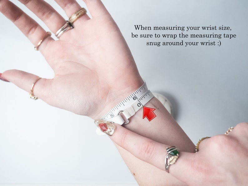 May include: A white measuring tape wrapped around a wrist, indicating a wrist size of approximately 6 inches. The image includes text advising to wrap the tape snugly. Several rings adorn the fingers.
